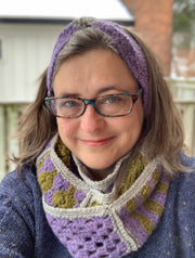 Image: middle aged white woman wearing glasses , blue sweater, lilac headband and crocheted infinity scarf in lilac, green and grey