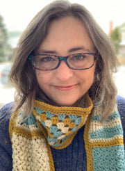 Image: middle aged white woman wearing glasses , blue sweater, lilac headband and crocheted infinity scarf in blue, ivory and gold